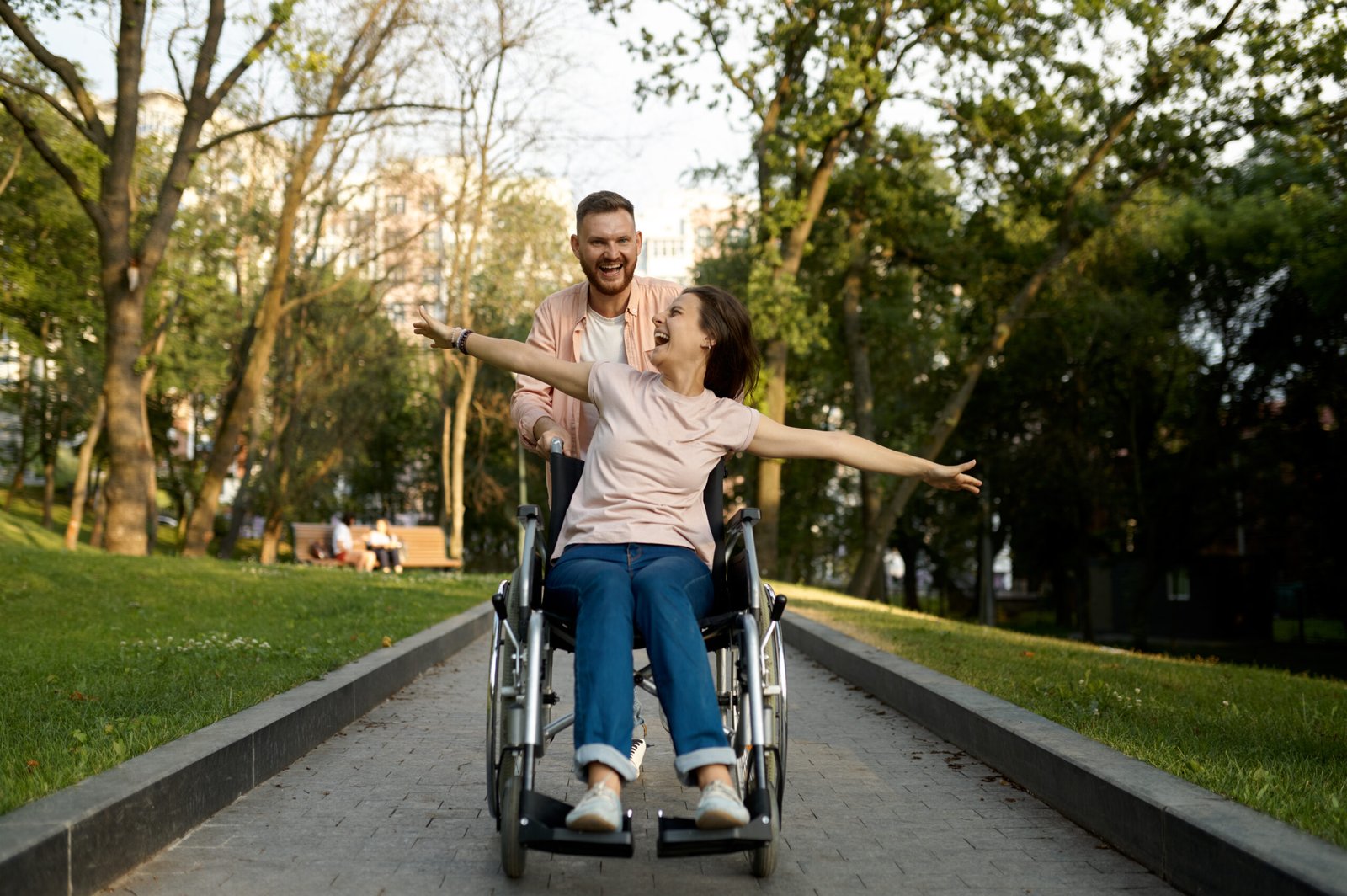 Man happily pushing a joyful woman in a wheelchair through a park on a sunny day.