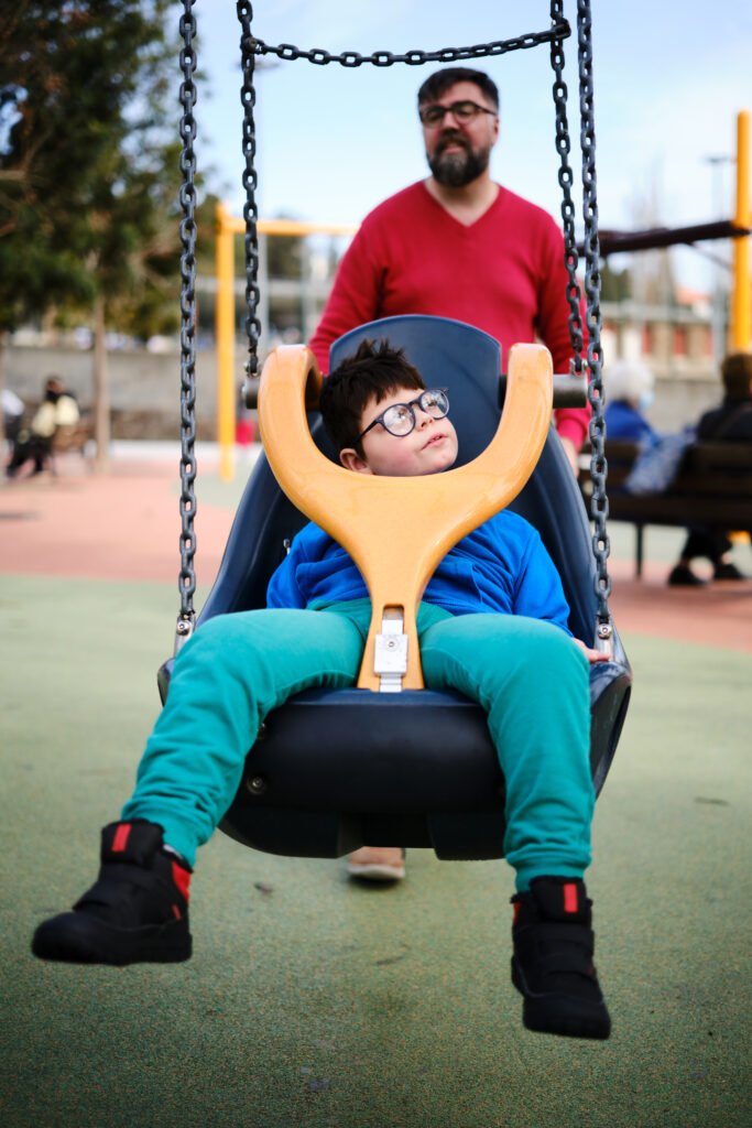 Young boy with disability on an adaptive swing at the playground, supervised by a carer.