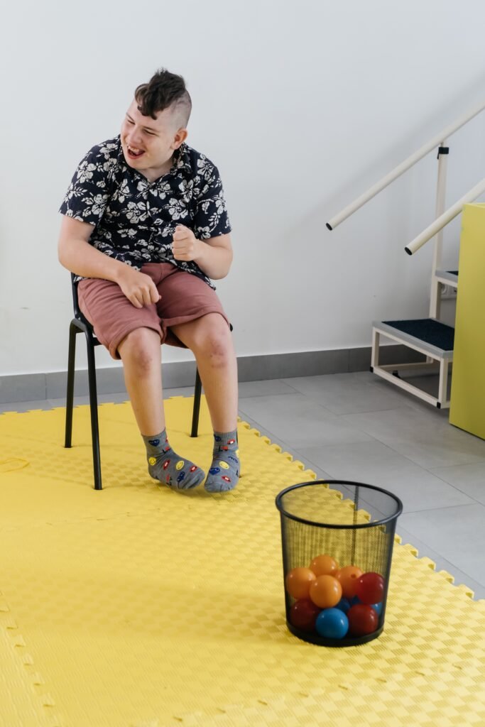Two NDIS participants smiling—one engaging in an indoor activity with balls, the other sitting outdoors beside a wheelchair—showcasing inclusive group programs at Improved Ability.Two NDIS participants smiling—one engaging in an indoor activity with balls, the other sitting outdoors beside a wheelchair—showcasing inclusive group programs at Improved Ability.