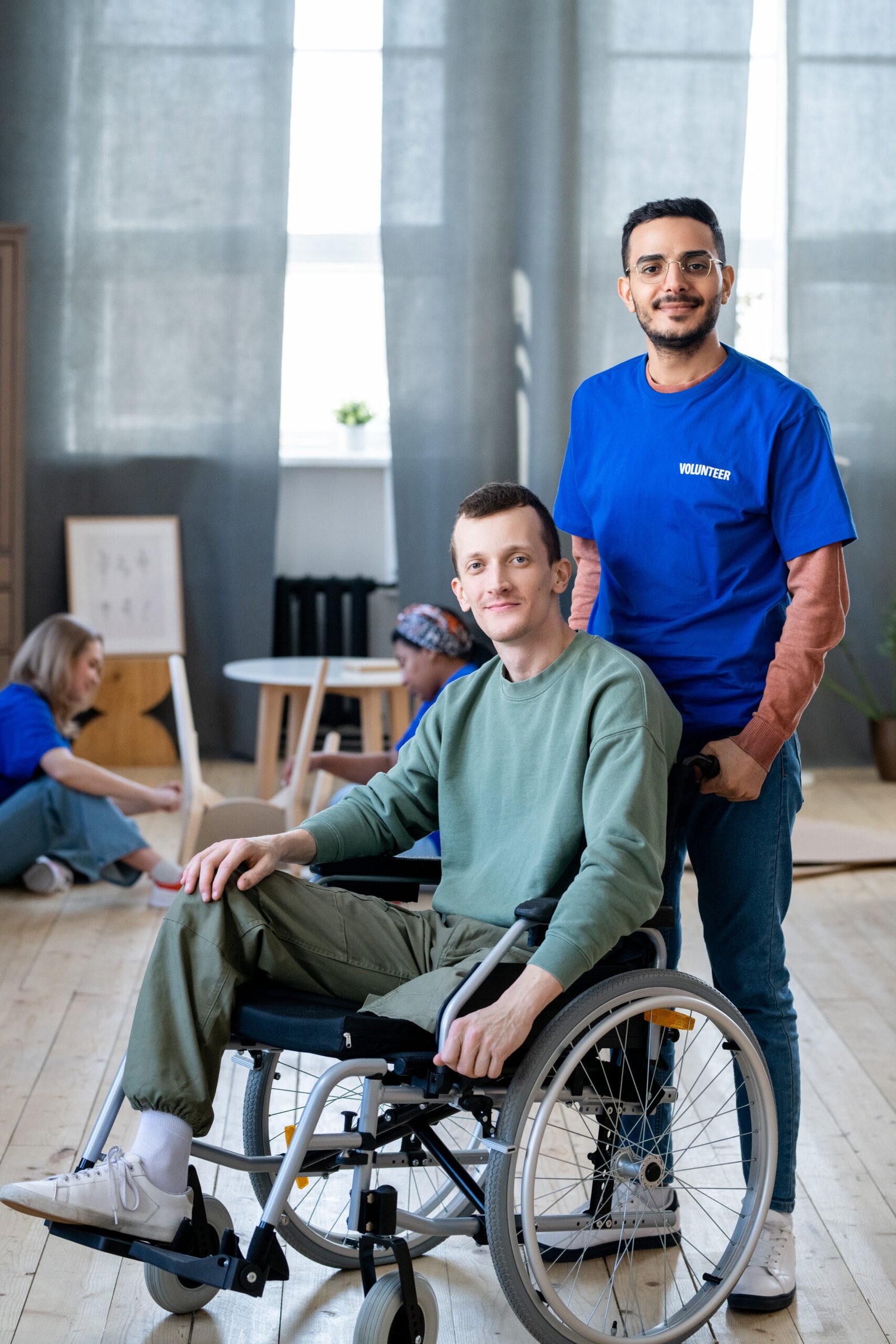Young man in a wheelchair with a volunteer standing beside him, smiling in a supportive community setting.